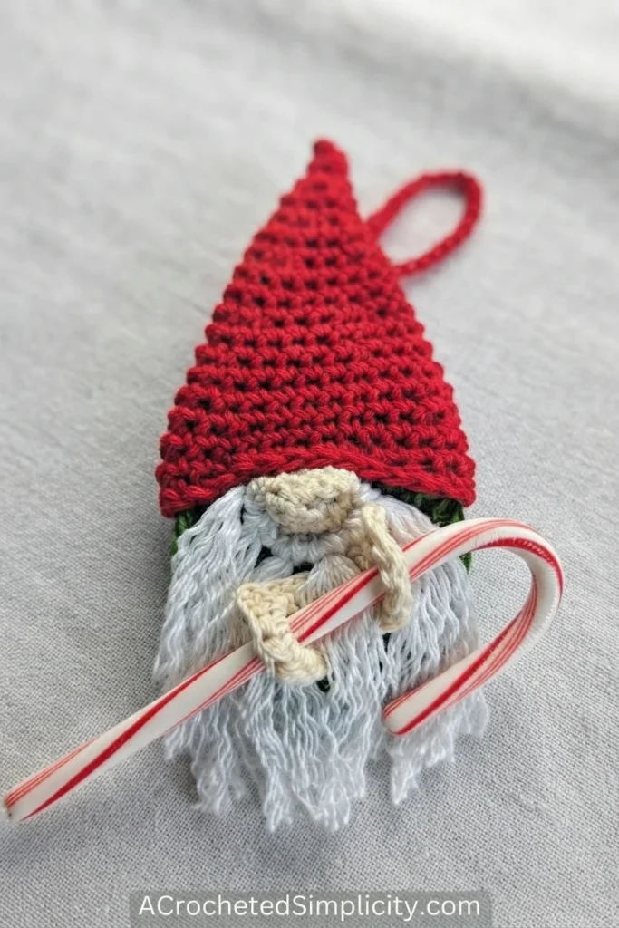 Crocheted gnome ornament with a red hat, white beard, hanging loop, and striped candy cane on gray fabric.