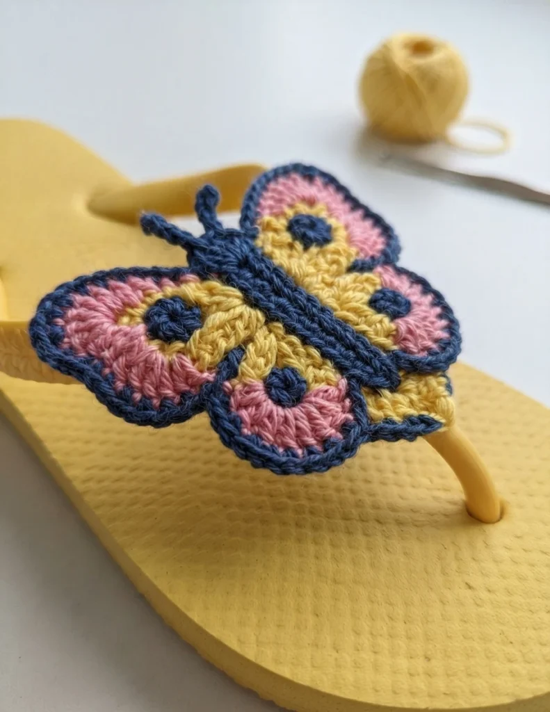 Yellow flip-flop with a crocheted butterfly motif in pink, yellow and dark blue, with a ball of yellow yarn and a crochet hook in the background