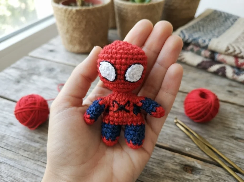 A small red-and-blue crocheted Spider-Man doll with white eyes is held in a hand above a wooden table, with yarn balls and a crochet hook nearby.