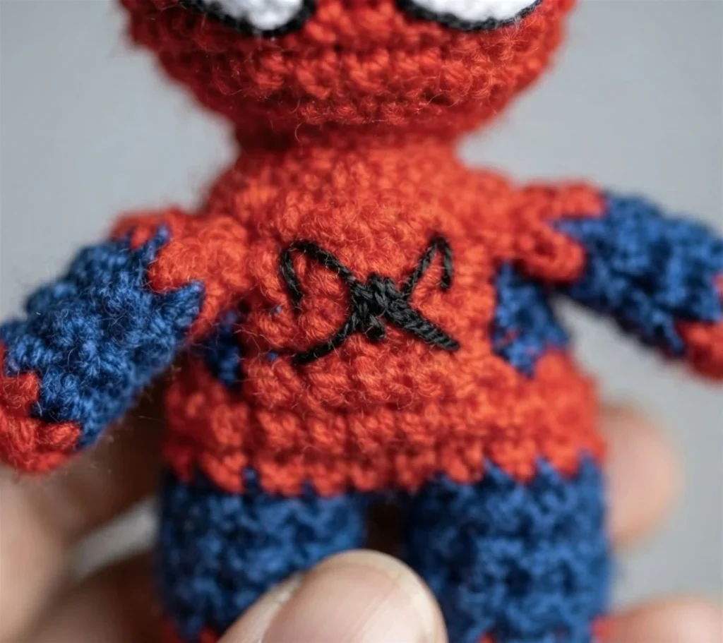 Close-up of a black spider emblem sewn onto the red chest of a red-and-blue crocheted doll.