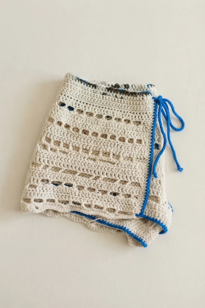 Vêtement au crochet écru à motifs ajourés, avec une bordure et des liens bleus, posé plié sur un fond clair.