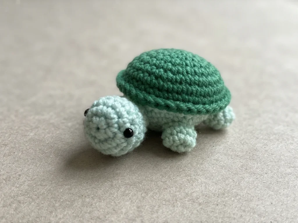 Small crocheted turtle with a dark green shell and pale mint head and legs on a light surface