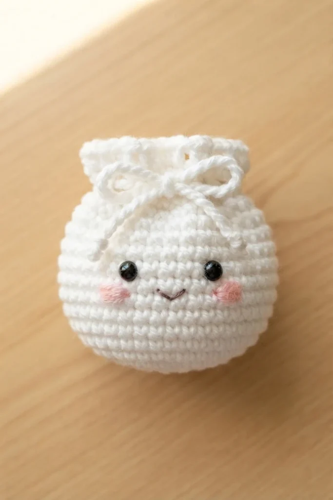 White crochet dumpling pouch with black safety eyes, pink cheeks and tied drawstring on a light wood surface