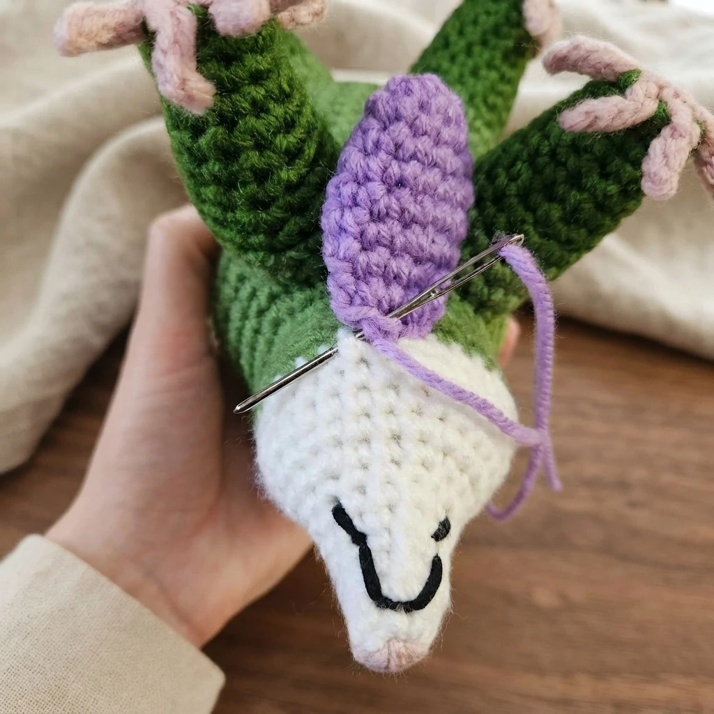 Crocheted opossum head in white and green held in one hand while a purple crocheted petal is sewn to the neck with a tapestry needle and purple yarn.
