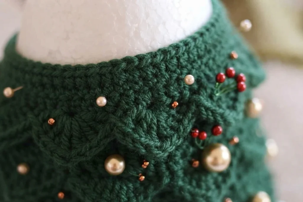 Close-up of a dark green crocheted ribbon wrapped around a white foam cone, secured with pearl-headed pins and decorated with gold beads and small red berries.