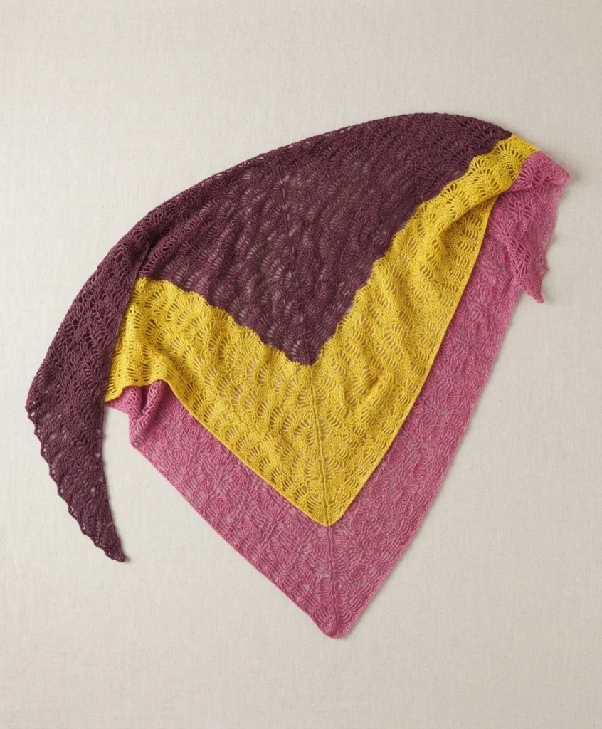 A folded lace shawl draped on a pale background, showing overlapping bands of plum, yellow and pink worked in an open fan stitch pattern.