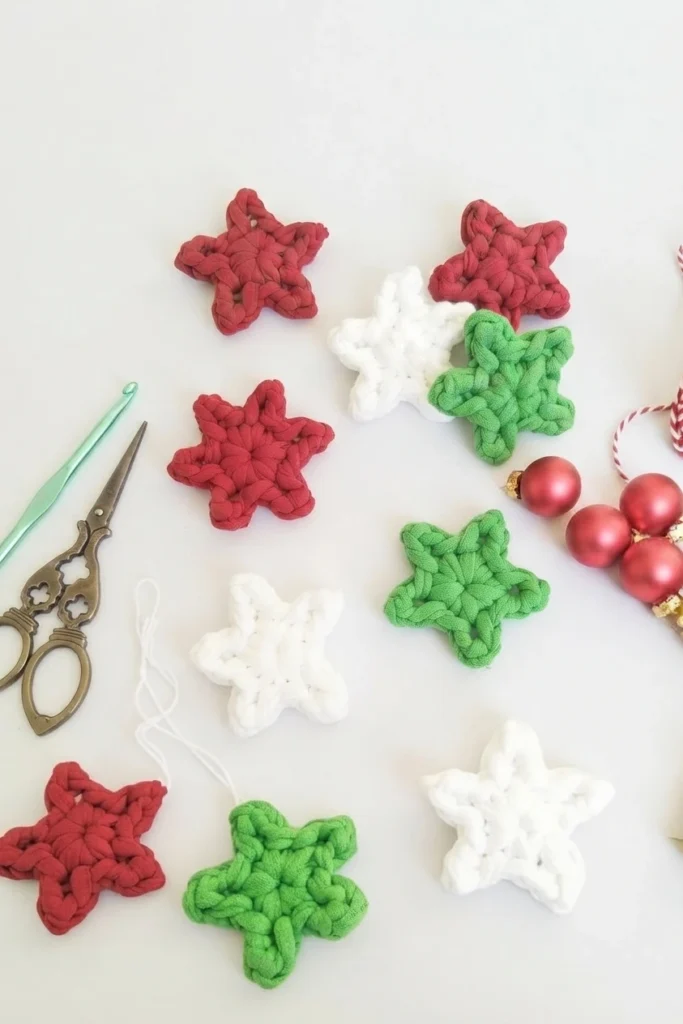 Several flat crocheted star ornaments in red, white and green t-shirt yarn arranged on a white surface, with vintage metal scissors, a green crochet hook and red Christmas baubles at the edge.