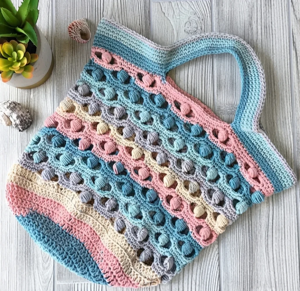 Crocheted market tote bag in blue, pink and cream stripes with raised shell bobble pattern lying on light wood beside a small potted plant and seashells