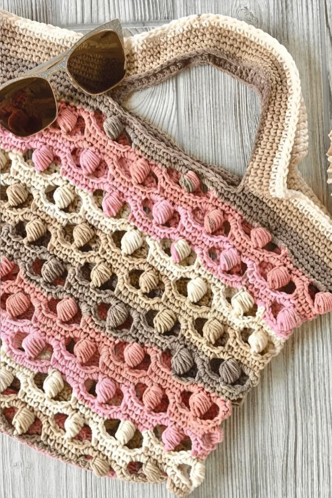 Crocheted market tote bag in pink, beige and brown with bobble shell motifs and sunglasses resting on top, on a light wooden surface