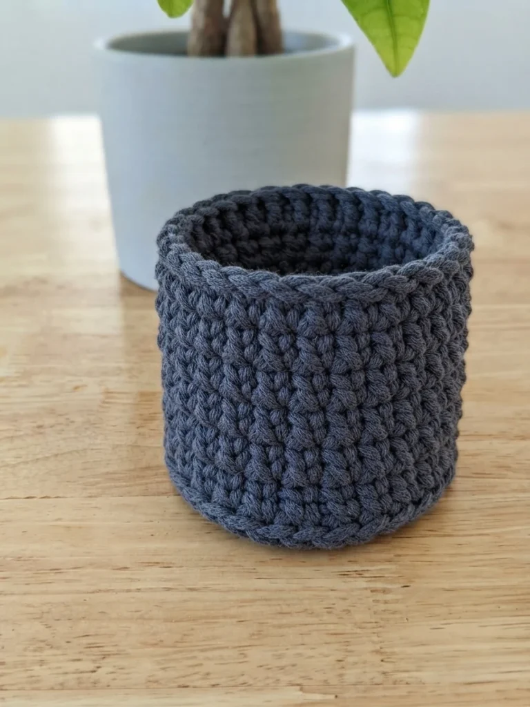 A blue-grey crocheted plant pot cover shown finished and empty on a wooden table, with a potted plant blurred behind it.