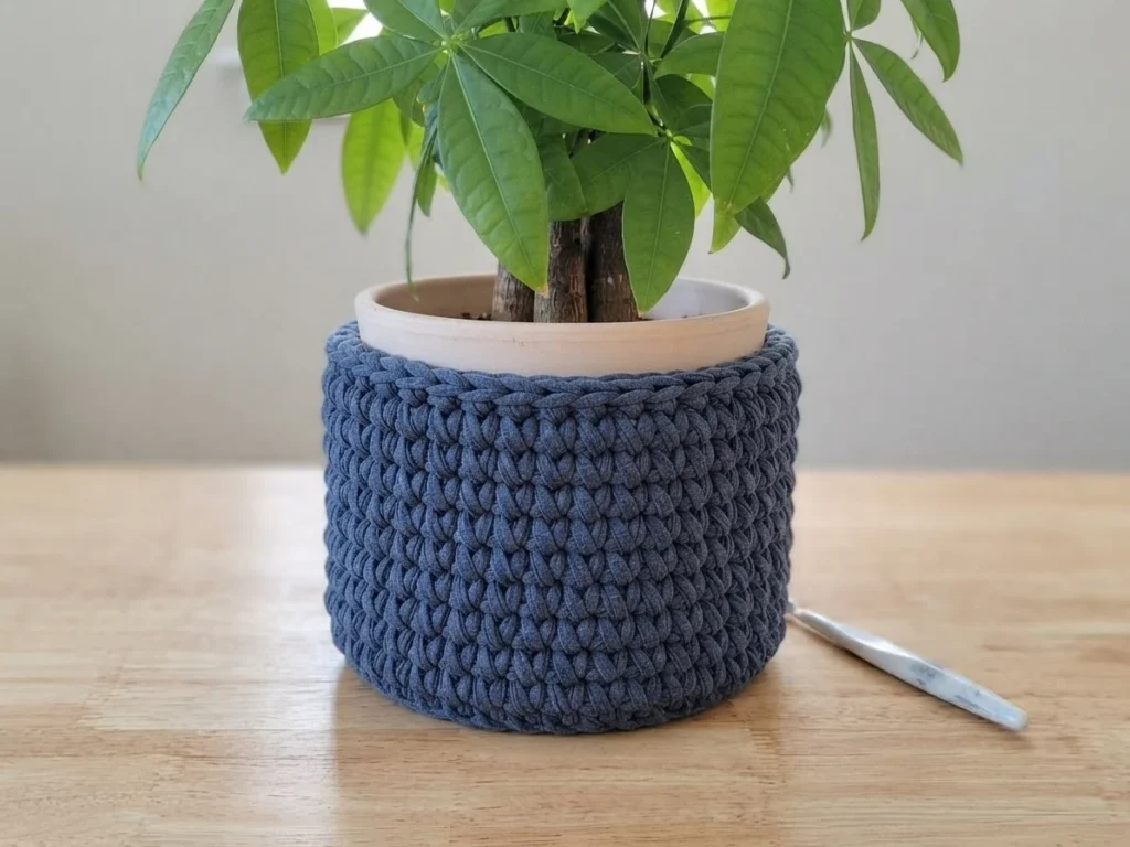 Dark blue crocheted plant pot cover around a potted green plant on a wooden table, with a crochet hook beside it.