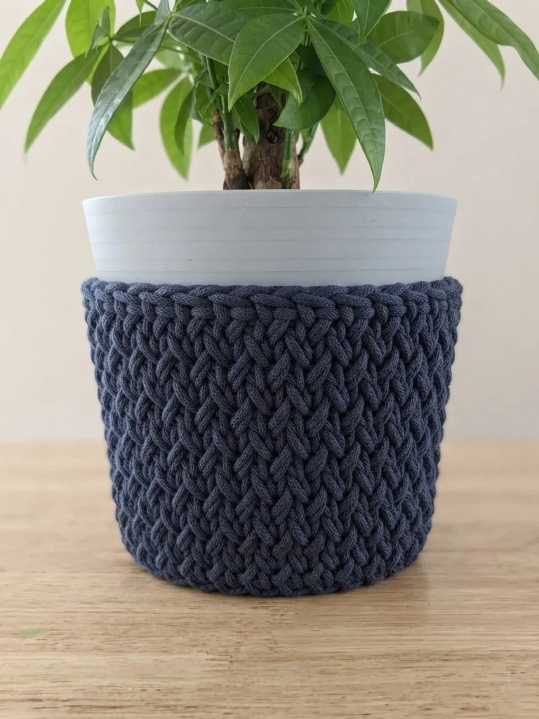 Dark blue crochet cover around the lower part of a white plant pot with a green leafy plant on a wooden table.