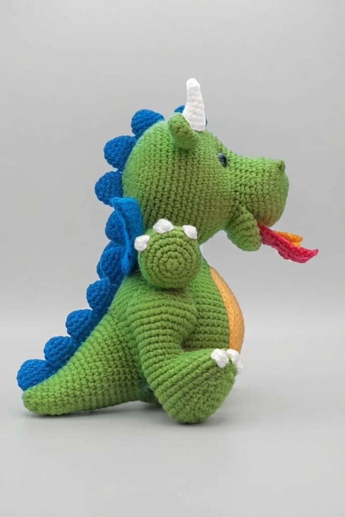Side view of a green crochet dragon with blue back spikes, white horn and claws, yellow belly patch and small red and orange flames at the mouth