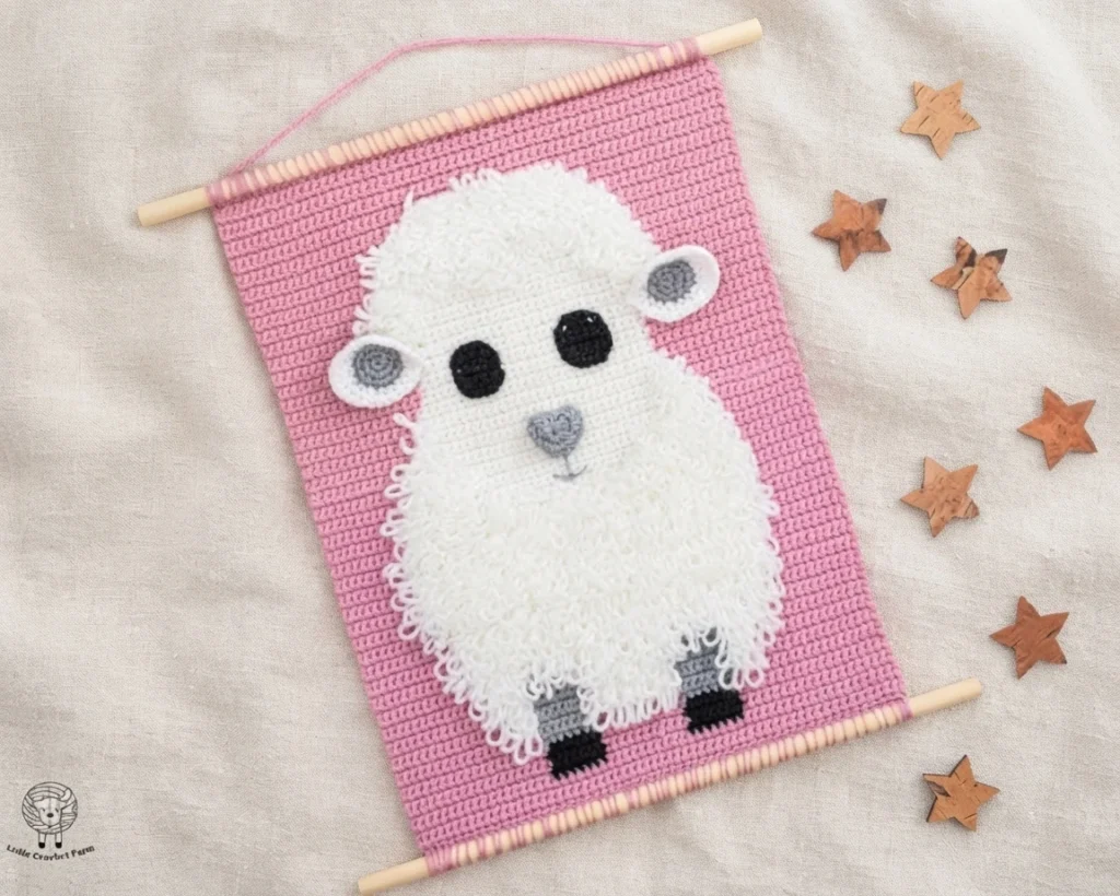 A pink crochet wall hanging with a fluffy white sheep, wooden dowels, and scattered star ornaments on beige fabric.