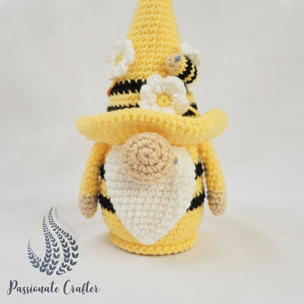 Finished crocheted bumble bee gnome with tall yellow striped hat, white beard, and a mini bee applique near the flower decorations.