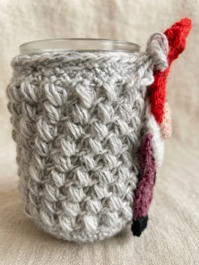 Mason jar wrapped in a grey textured crochet cover with a small gnome appliqué on the side, featuring a red hat, pink nose, white beard, plum body and black boots.