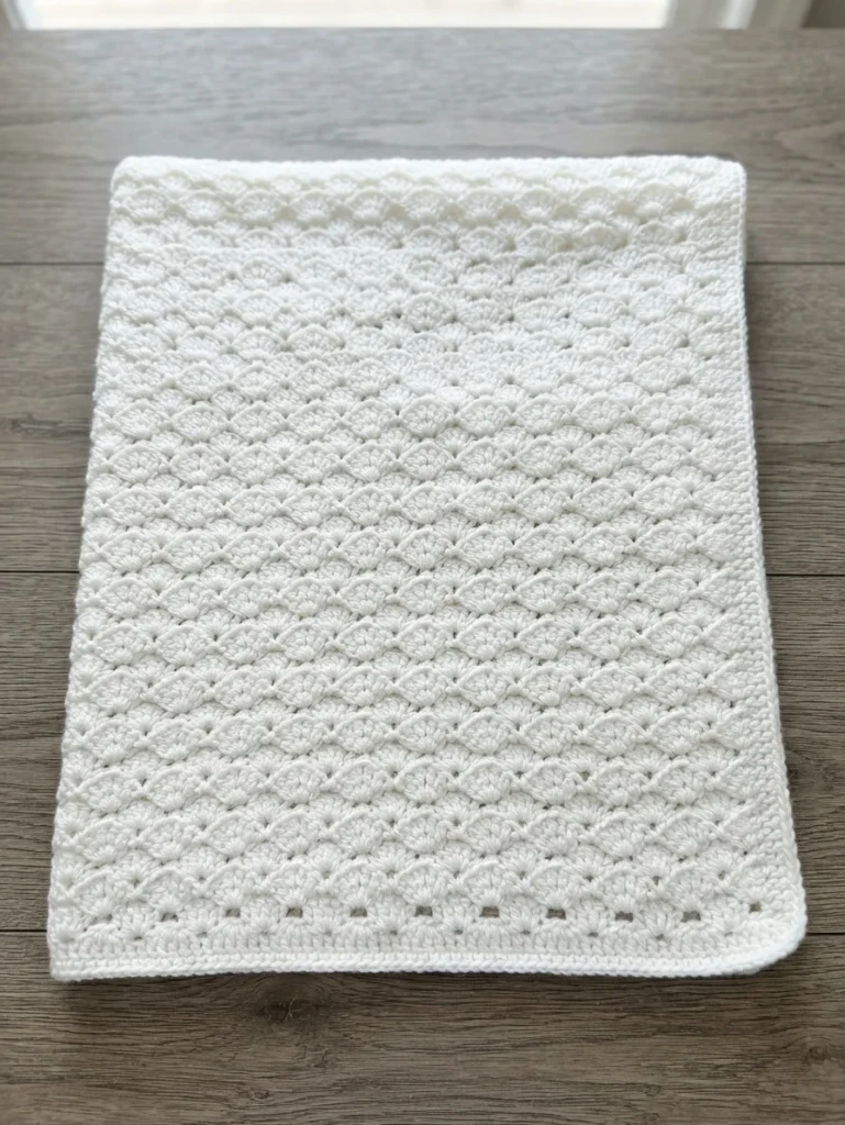White crocheted baby blanket with a shell stitch pattern laid flat on a wooden table.
