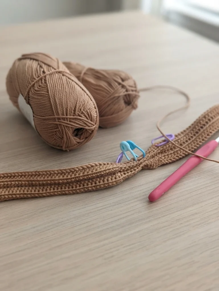 Two balls of tan yarn with a crocheted strip in progress, stitch markers and a pink crochet hook on a light wooden table.