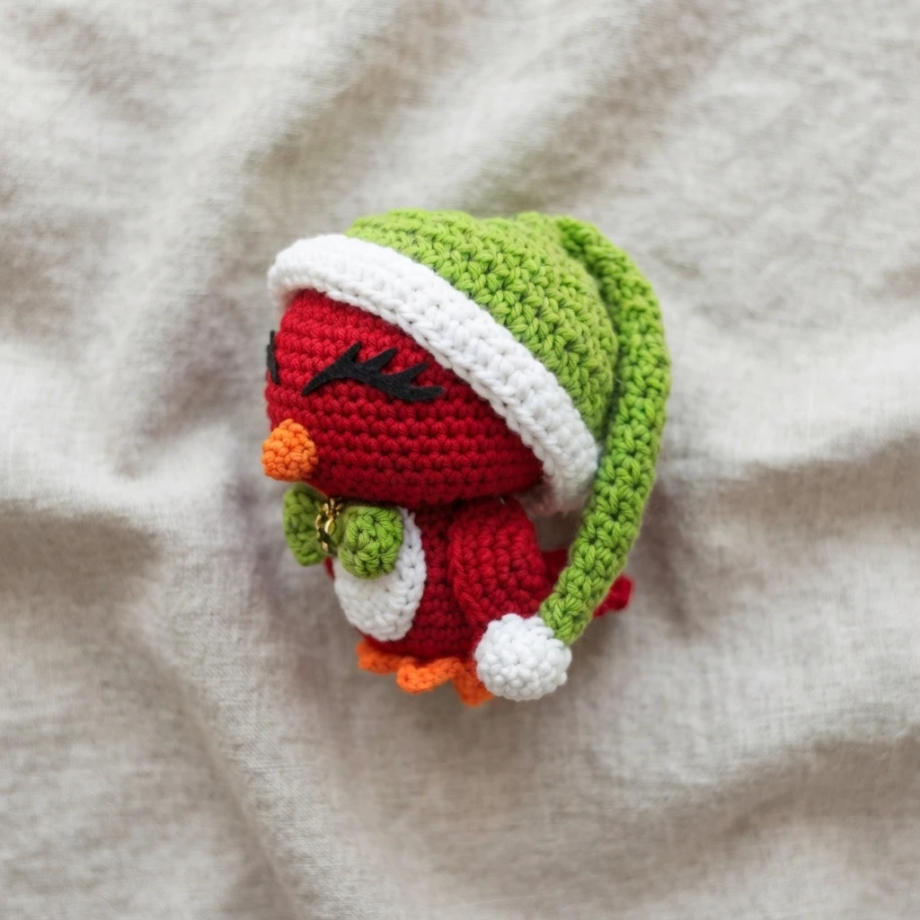 Crocheted red cardinal ornament wearing a green Santa hat with a white brim and white pom-pom, with an orange beak, black stitched brow, green wings and orange feet, resting on a light beige fabric.