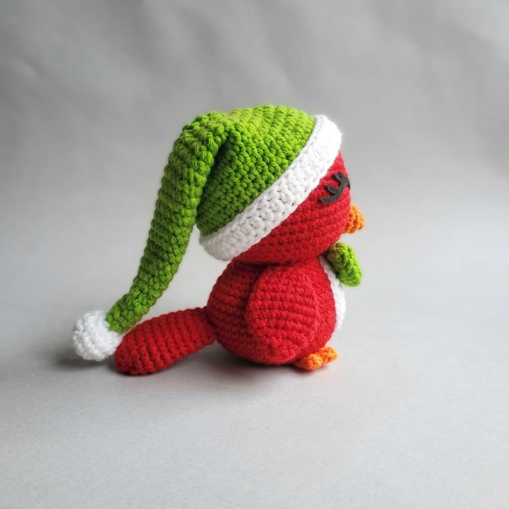 Crocheted red cardinal bird with a green Santa hat trimmed in white, orange beak and orange feet, shown from the side on a plain gray background.