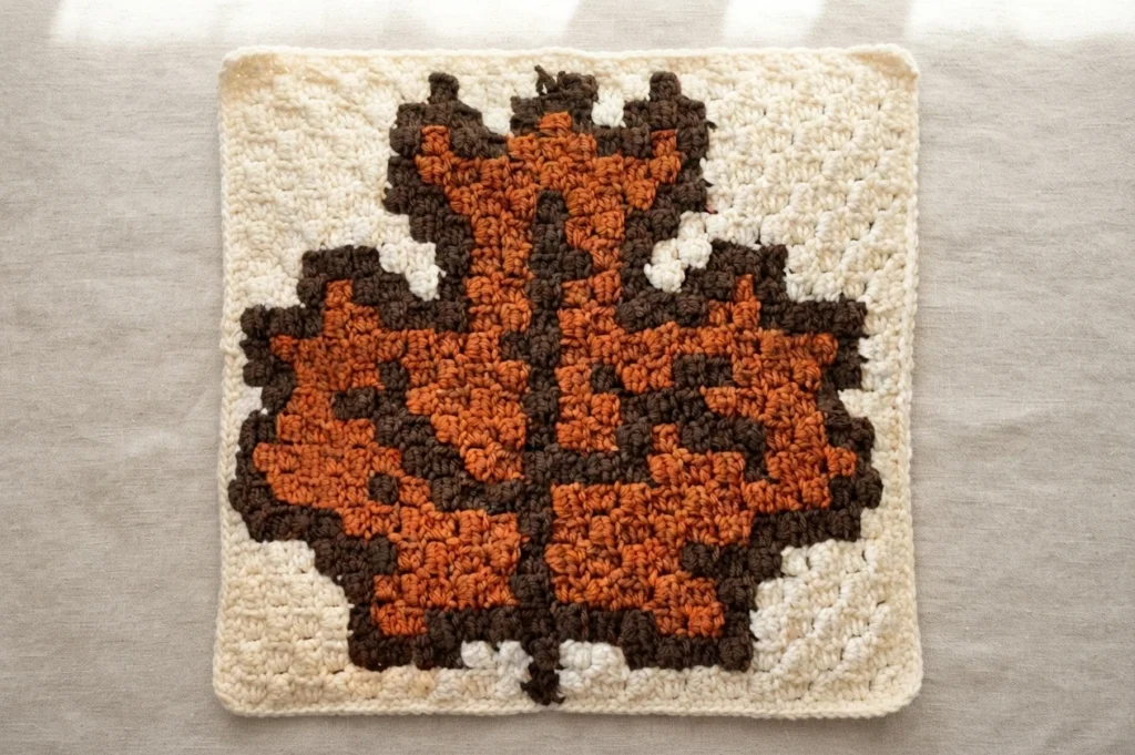 Crochet square worked in corner-to-corner stitch showing an autumn maple leaf in orange and brown on a cream background, laid flat on a plain light surface.