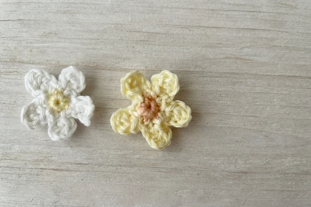 Two small crocheted daisies, one white with a pale yellow center and one pale yellow with a peach center, on a light wood surface.