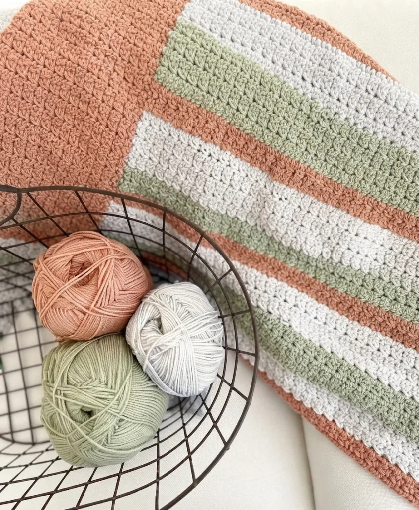A cream, sage green, and terracotta crocheted blanket draped over a chair beside three yarn balls in a wire basket.