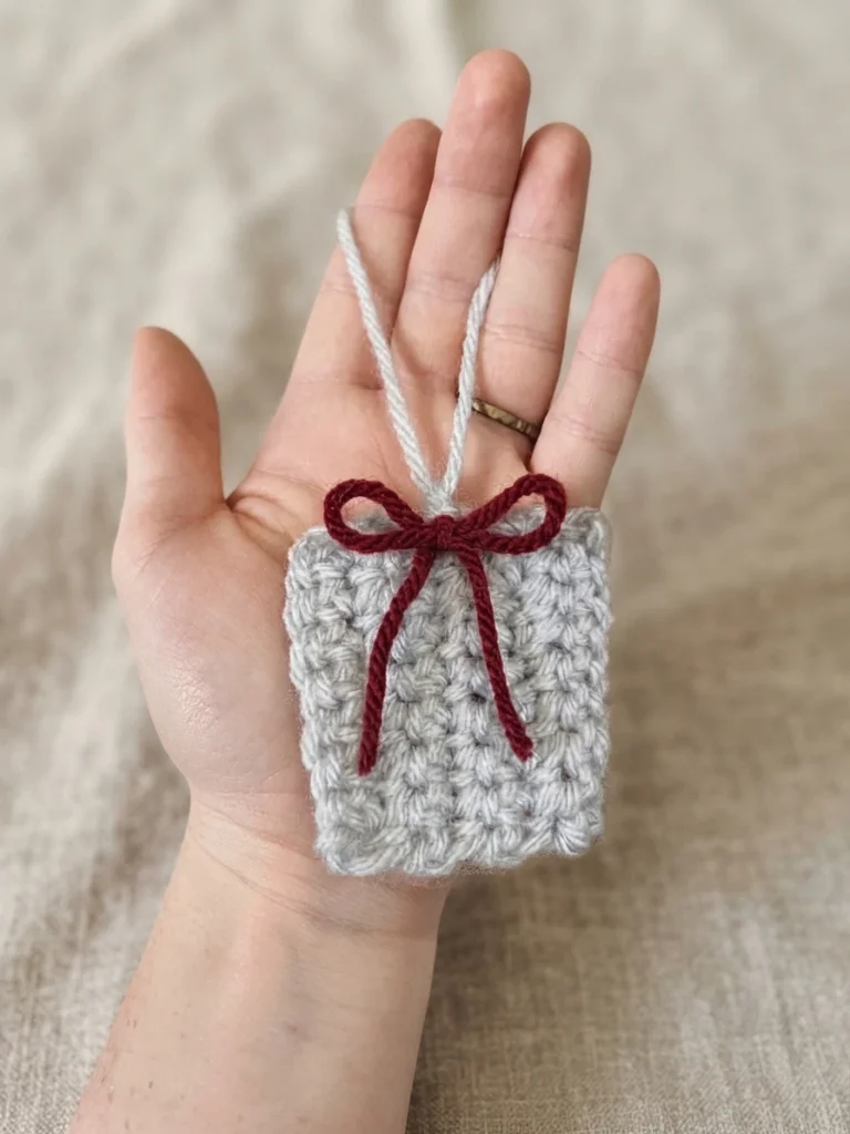 A small gray crocheted gift ornament with a burgundy bow and hanging loop, held in an open hand.