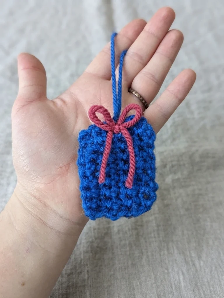 A hand holds a small blue crocheted gift ornament with a pink bow and a blue hanging loop.