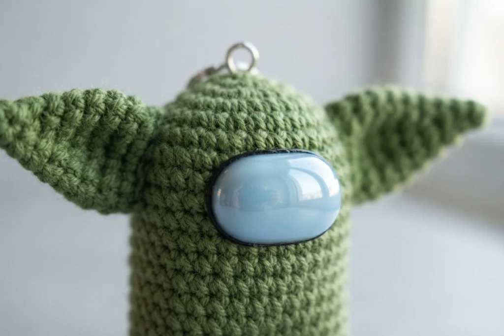 Close-up of a green crocheted crewmate keychain head with long pointed ears and a pale blue visor.