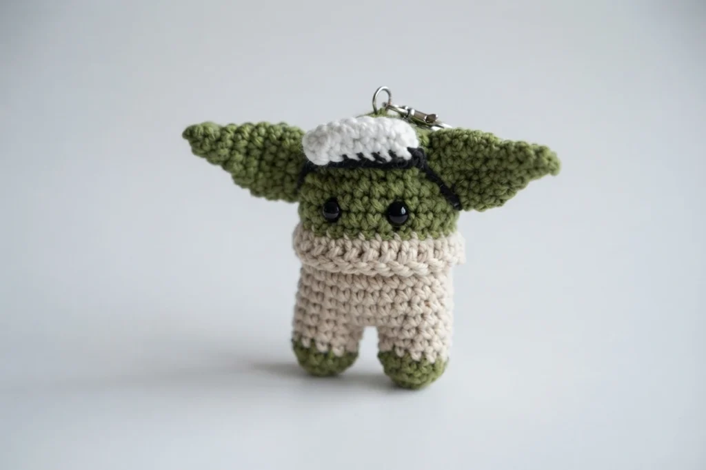 Crocheted green Baby Yoda Among Us crewmate keychain with large ears, beige body, safety eyes and a white patch on its head.