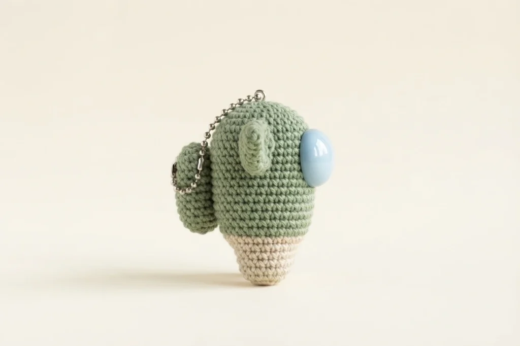 Side view of a crocheted Baby Yoda Among Us crewmate keychain in sage green and beige with a light blue visor and ball chain, on a plain background.