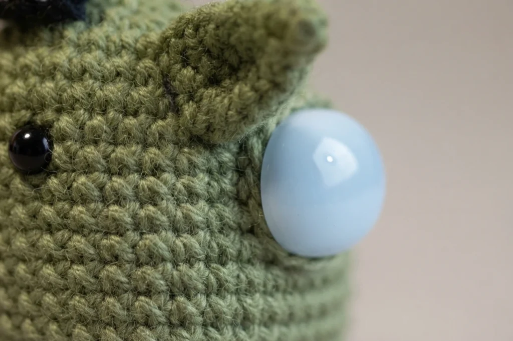 Extreme close-up of a green crocheted Baby Yoda Among Us crewmate head showing textured stitches, one pointed ear, a black safety eye and a large pale blue oval shape on the side