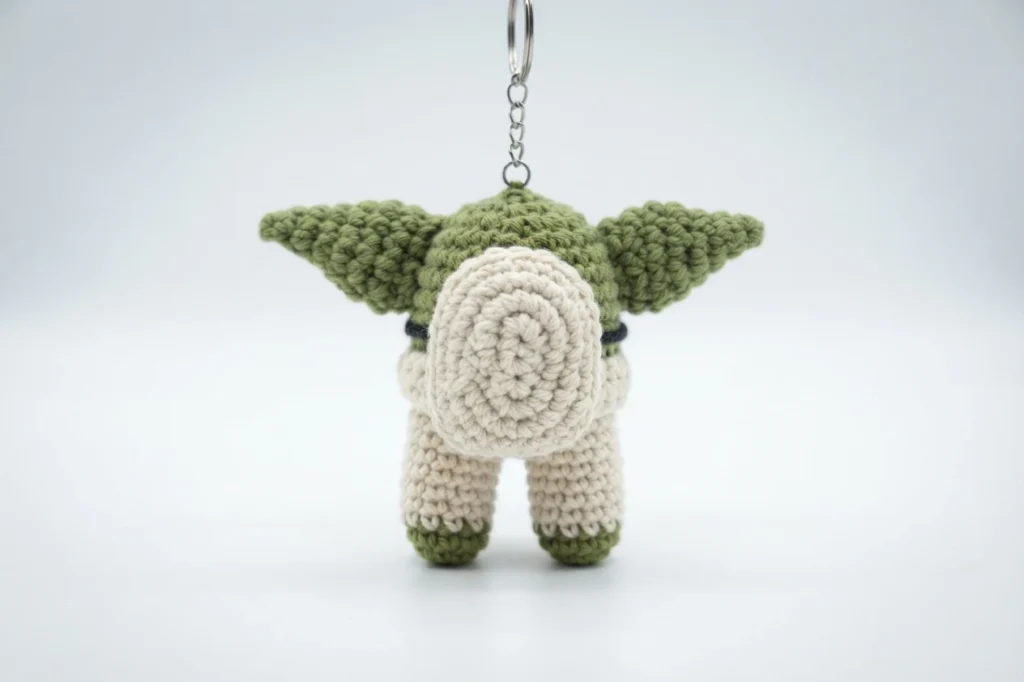 Back view of a crocheted Baby Yoda Among Us crewmate keychain with a green head, large pointed ears, cream backpack, beige legs and green feet, hanging from a metal keyring on a white background.