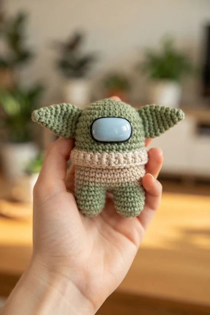 A hand holding the finished crocheted green crewmate figure with pointed ears, a light blue visor and a beige robe.