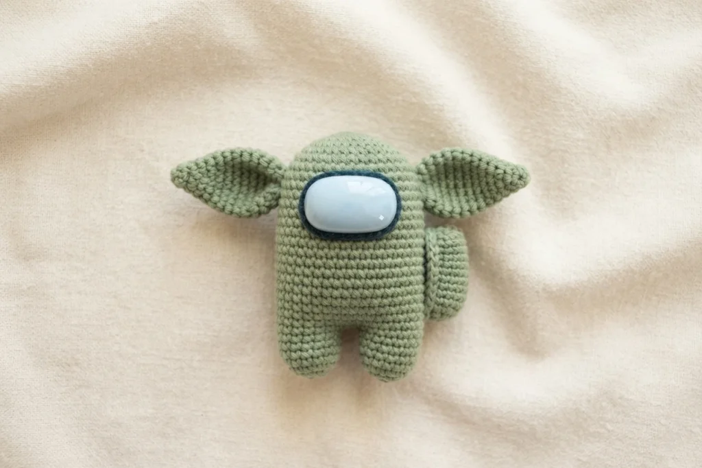 Small green crocheted crewmate figure with large pointed ears and a light blue visor resting on cream fabric