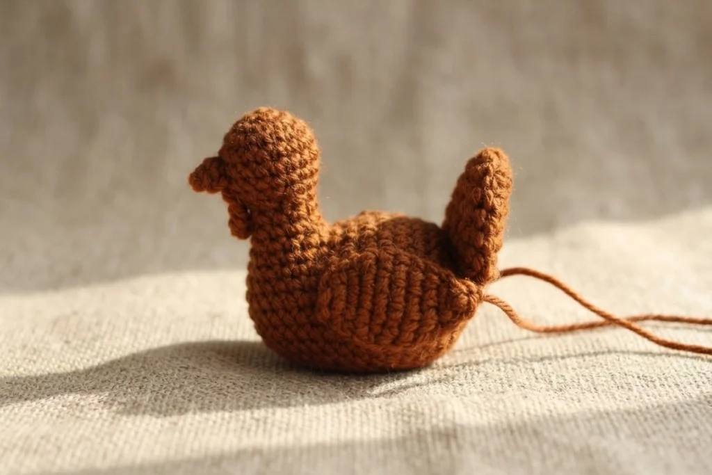 Brown crocheted amigurumi turkey with a rounded body, raised head and small beak, and an upright tail feather, sitting on beige linen fabric with loose yarn ends