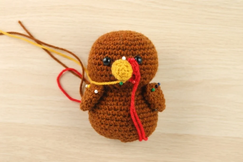 Crocheted brown turkey amigurumi in progress, with a yellow beak, red wattle, black safety eyes, small colored pins marking placement and loose yellow, red and brown yarn ends, resting on a light wood surface.