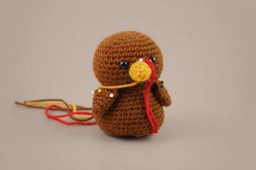 Crocheted brown turkey amigurumi with black safety eyes, a yellow beak and a red wattle, its wings and face pieces pinned in place with a yellow thread and loose red and yellow yarn ends resting on a beige surface.