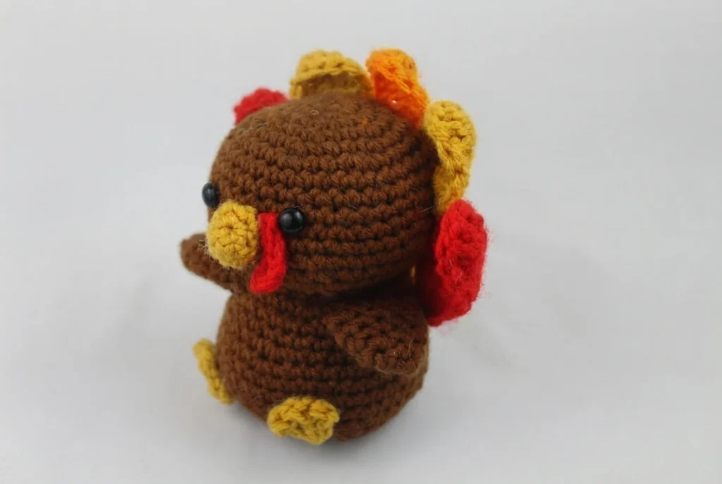 Crocheted brown turkey amigurumi with black safety eyes, a yellow beak, a red wattle, small yellow feet and red, orange and yellow tail feathers, photographed on a plain white surface.