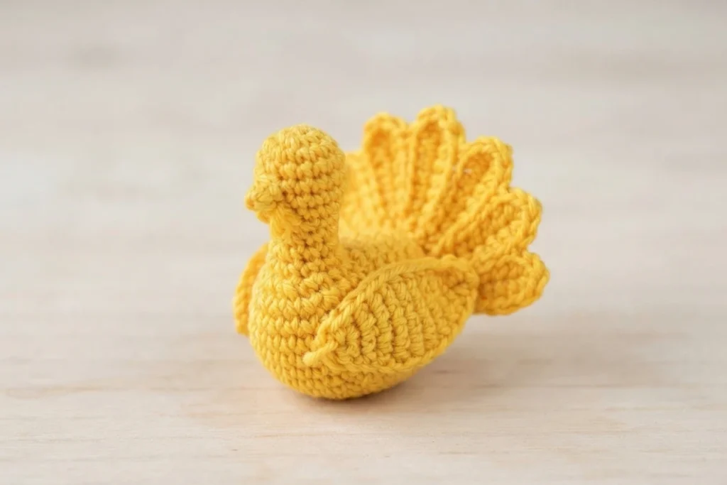 Yellow crocheted turkey amigurumi with a rounded body, small head, and a fan of tail feathers, sitting on a light wood surface.