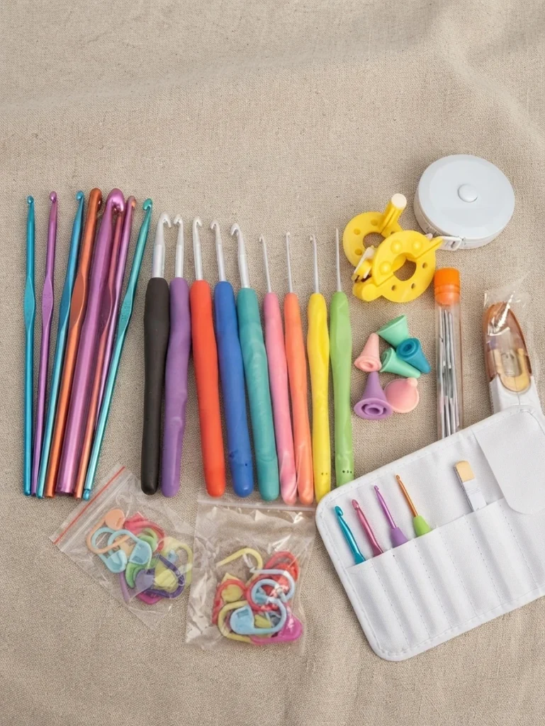 Crochets de tailles et de couleurs variées, accessoires de crochet et étui blanc disposés sur un tissu beige.