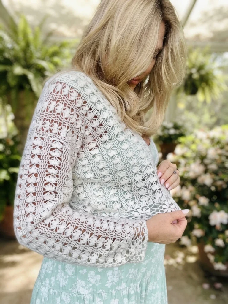 Woman with long blonde hair modelling a white openwork crochet cardigan with flower motifs over a mint green floral dress, standing among potted plants and roses in a greenhouse