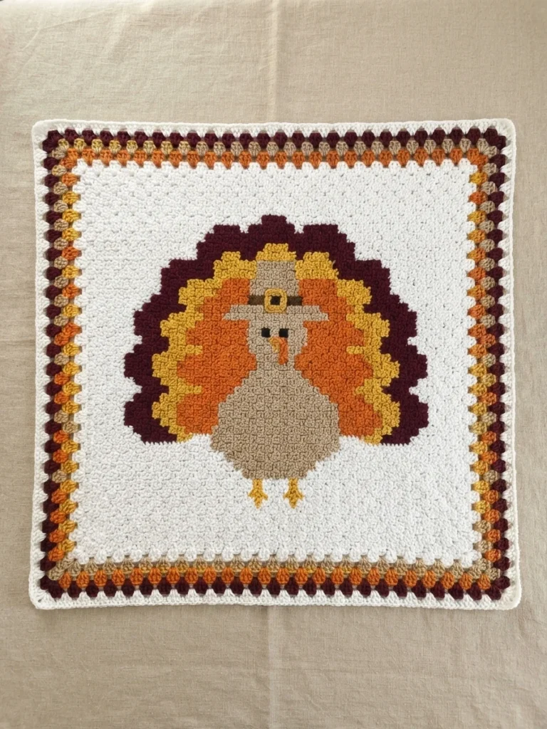 Crochet corner to corner turkey blanket on a white background, centered turkey with a pilgrim hat, small dark eyes and orange beak, fanned tail feathers in dark maroon, orange, yellow and tan, surrounded by striped block stitch border round