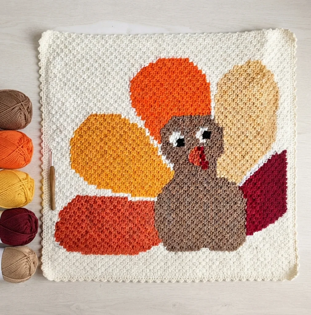 Square crochet blanket with a brown turkey in the center, its tail feathers fanning out in orange, yellow, tan and dark red on an off-white background, laid flat on a pale wood surface beside five balls of yarn and a wooden crochet hook.