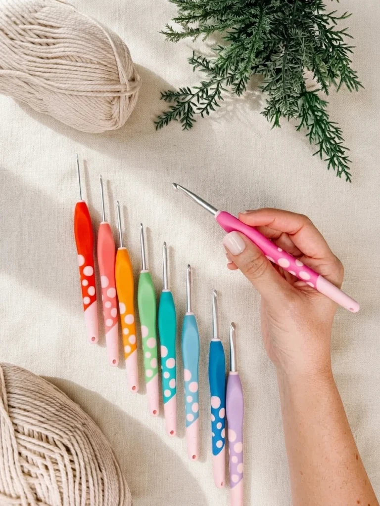 A hand holds a pink crochet hook beside a row of colorful crochet hooks, beige yarn, and greenery.