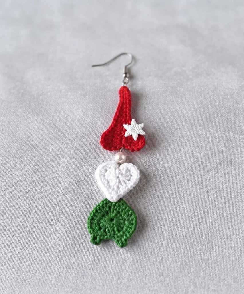 Crocheted gnome earring hanging from a silver ear wire, with a red heart-shaped cap topped by a small white snowflake button, a pearl bead below it, a white crocheted heart beard and a green crocheted heart body with two small feet, photogr