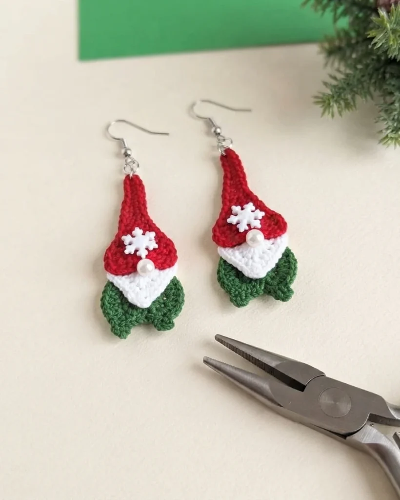 Pair of crochet gnome earrings in red, white and green with white snowflake buttons, pearl noses and silver ear wires, laid on a pale surface beside flat nose pliers and greenery.
