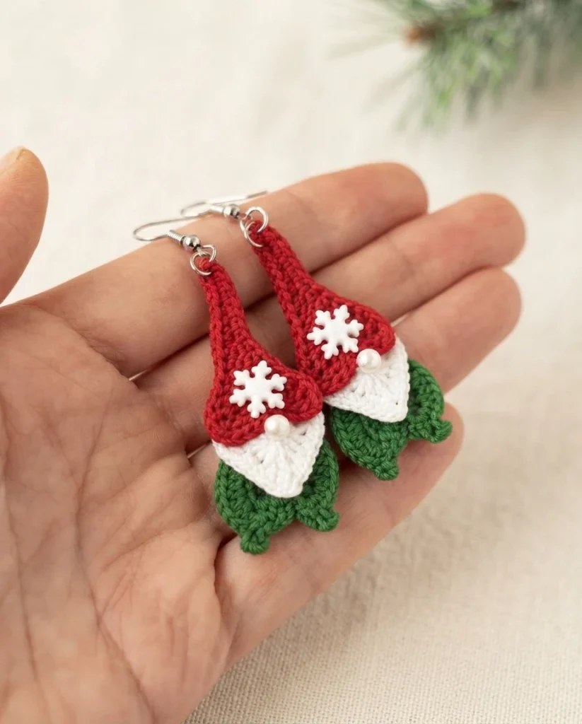 Pair of crocheted Christmas gnome earrings in red, white and green held in an open hand, each with a white snowflake button on the hat, a pearl bead nose and a silver ear wire.