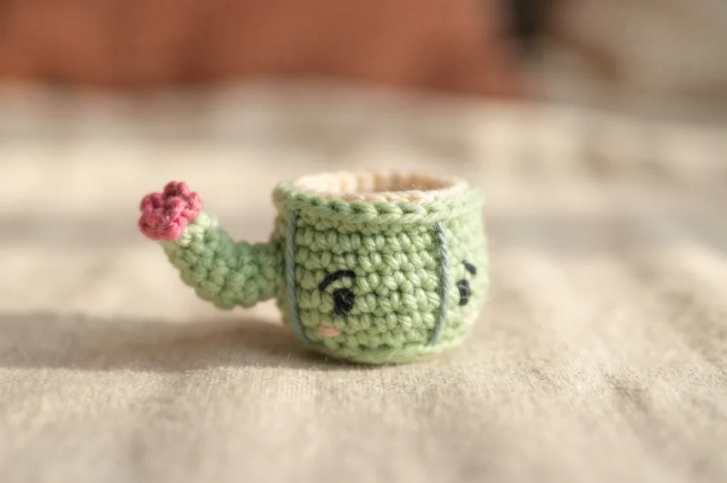 Crochet mini cactus mug amigurumi with a small arm tipped with a pink flower and an embroidered face.